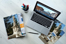 Laptop displaying a scenic image on a desk with a magazine, pen, and postcard.